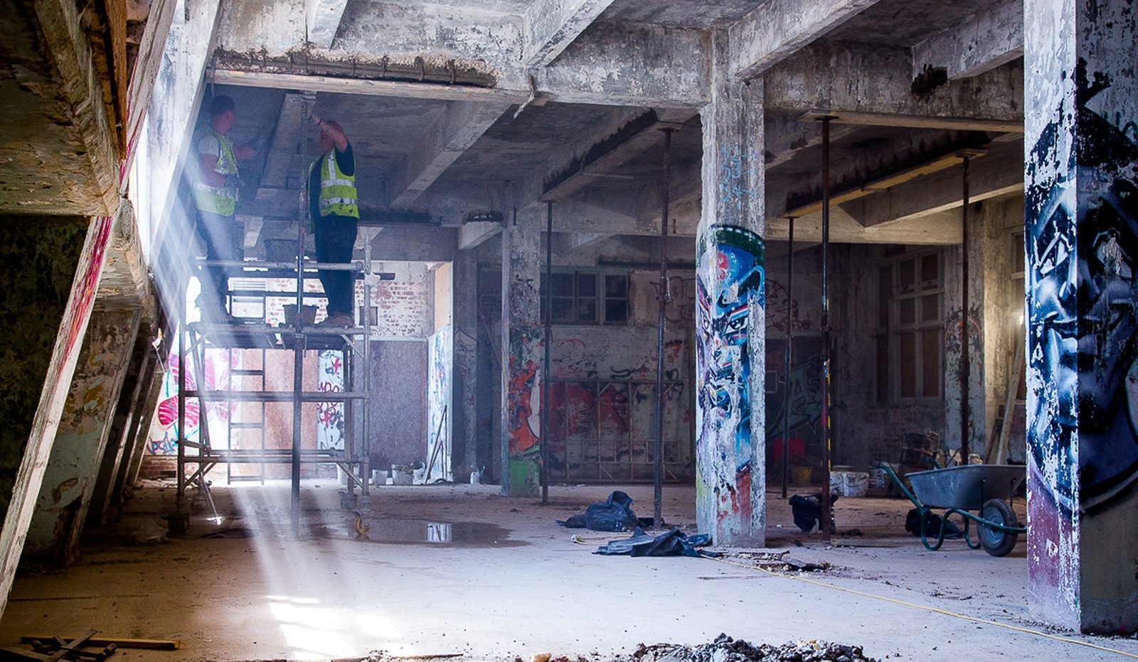 Derelict warehouse interior with light streaming through, two workers on scaffolding